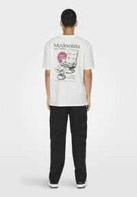 T-shirt blanc avec un motif au dos représentant un design de chaise des années 50, texte noir et accents roses. Pantalons cargo noirs et baskets blanches visibles.