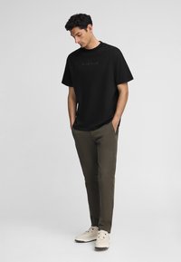 Camiseta negra con escote redondo y mangas cortas, combinada con pantalones verde oliva. El conjunto se completa con zapatillas de deporte en color blanco sucio.