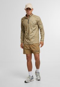 Beige Langarmshirt mit geometrischem Muster und Reißverschlusskragen, kombiniert mit beigen Shorts mit einem dunkleren Muster und Logo, ergänzt durch Laufschuhe.