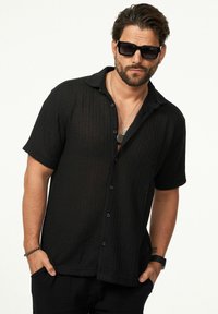 Schwarzes Kurzarm-Hemd mit Knopfleiste aus strukturiertem Stoff und lockerer Passform, kombiniert mit dunklen Sonnenbrillen und geschichteten Halsketten.