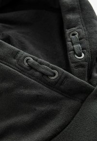 Grembiule nero con cappuccio realizzato in tessuto testurizzato, dotato di cordoncino e occhielli in metallo. Il design include un orlo liscio e piatto.