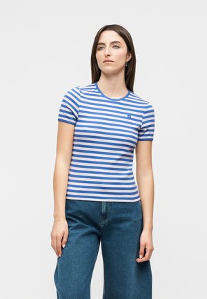 Γυναίκα που φοράει μπλε και λευκή ριγωτή μπλούζα με κοντά μανίκια και μπλε τζιν, στέκεται μπροστά σε ένα απλό φωτεινό φόντο.