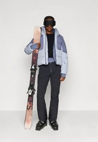 Slēpošanas tērps ar gaismzilas un pelēkas krāsas pūkaino jaku, melnu pamatslāni, melnas bikses un melnus slēpošanas zābakus, turot slēpes ar stiprinājumiem.