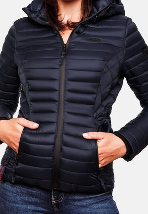 Person iført en navyblå quiltet jakke med lynlås med hænder i sidelommerne og blå jeans, vist fra nakke til hofter.