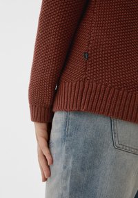 Gestrickter Pullover in Terrakotta, mit strukturiertem Muster und geripptem Saum. Kombiniert mit hellblauer Denim, zeigt eine Seitenansicht der Hand.