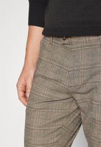 Man in beige geruite broek en een zwart gebreid sweater, met een hand ontspannen naast het been.