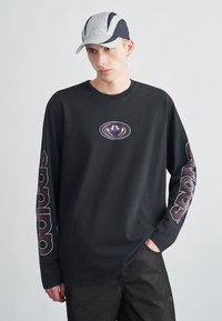 Juoda ilgomis rankovėmis marškinėliai su violetiniu logotipu ant krūtinės ir baltais užrašais ant rankovių, derinami su pilka ir juoda kepure su logotipu.