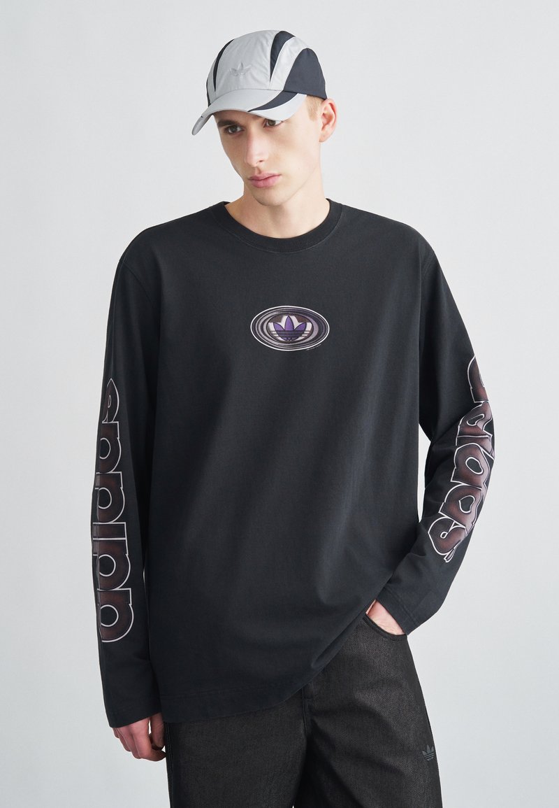 Juoda ilgomis rankovėmis marškinėliai su violetiniu logotipu ant krūtinės ir baltais užrašais ant rankovių, derinami su pilka ir juoda kepure su logotipu.