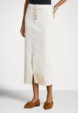 Naine kannab valget kõrge vöökohaga midi-seelikut nööpidega ees ja eesmist lõhikut ning pruune seemisnahast mokassiine tuttkestega.