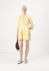 Vestido amarillo con detalles de encaje, mangas tres cuartos y falda de capas. La modelo sostiene un bolso oscuro y lleva zapatos de color claro.