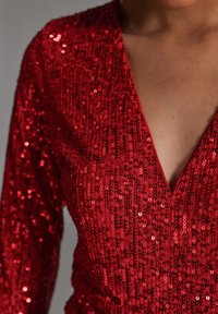 Rode pailletten jurk met een diepe V-halslijn en lange mouwen. Heeft een gestructureerd, glanzend oppervlak met een aansluitend ontwerp.