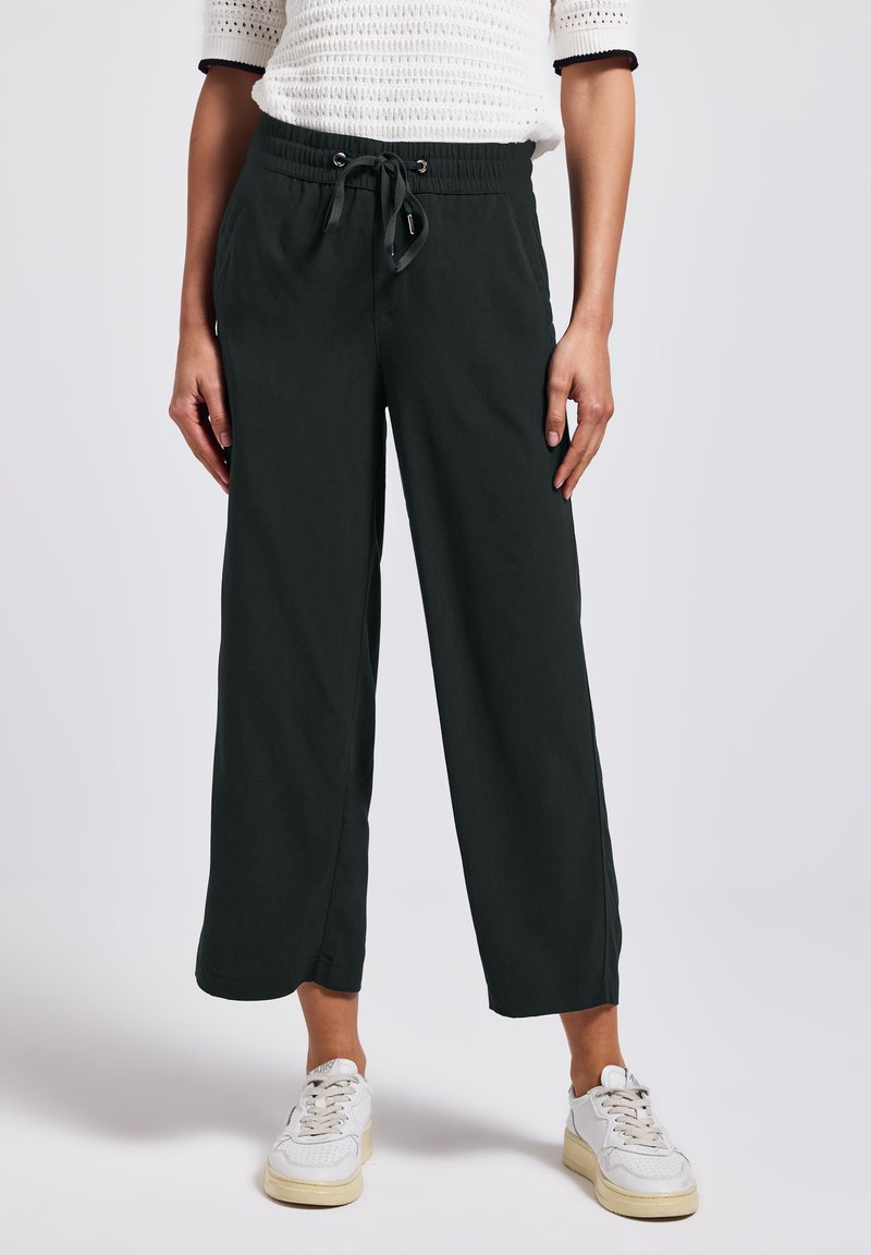 Černé kalhoty s širokými nohavicemi, elastickým pasem a šňůrkou, s hladkou texturou látky a bočními kapsami. V páru s bílými teniskami.