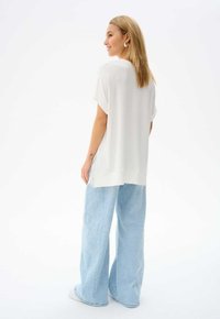 Witte, ruimvallende top met korte mouwen en zijsplitten, gecombineerd met lichtblauwe wijde jeans en witte sneakers. Minimalistisch ontwerp.