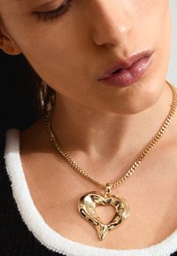Collier en pendentif en cœur doré avec un design texturé, suspendu à une chaîne épaisse et torsadée. Le pendentif présente une forme de cœur ouvert.