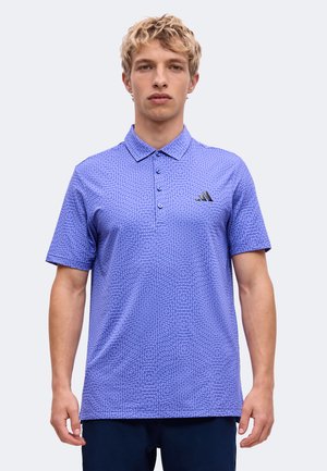 Jeune homme aux cheveux blonds portant un polo bleu à manches courtes avec un motif, trois boutons et un petit logo sur la poitrine.