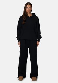 Zwarte oversized hoodie met een buidelzak en geborduurd logo, gecombineerd met tapered zwarte joggers en bruine instappers.