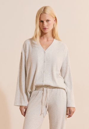 Donna bionda che indossa un top bianco di maglia a coste, ampio, con bottoni e pantaloni coordinati con coulisse, su sfondo beige semplice.