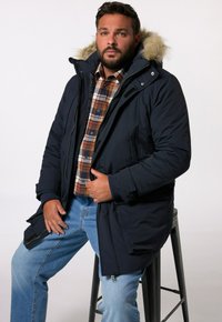 Marinblå parkas med foder av päls, dragkedja fram, och sidofickor, lagd över en rutig skjorta och kombinerad med ljusblå jeans.