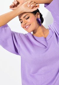 Maglione lavorato a maglia lavanda con maniche a palloncino a coste; il tessuto sembra morbido e leggero. Notevoli orecchini viola statement completano il look.