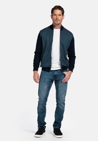 Blauwe bomberjack met donkerblauwe mouwen, wit T-shirt eronder, gecombineerd met blauwe jeans en zwarte sneakers. Voorzijde met rits.