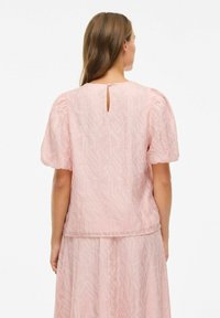 Lyserød bluse med pufærmer, en åbning i ryggen og struktureret mønster med broderede detaljer. Blødt, gennemsigtigt stof.