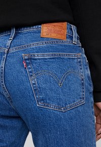 Jeans de mezclilla azul con un parche de cuero en la cinturilla y un diseño de bolsillo trasero distintivo que presenta un patrón de costura curvada.