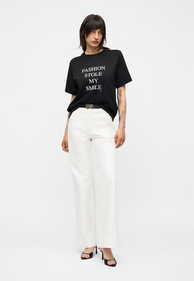 Γυναίκα με σκούρα μαλλιά που φοράει μαύρο μπλουζάκι με το κείμενο «FASHION STOLE MY SMILE», λευκό φαρδύ παντελόνι, μαύρα σανδάλια με τακούνι και τατουάζ στα χέρια.