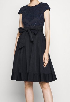 Vrouw draagt een knielange zwarte jurk met kant- en paillettendetails op het lijfje en een brede stoffen ceintuur met strik in de taille.