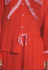 Punane pluus volangide ja roosade aktsentidega, nööpidega kinnitatav ja vöökohal seotav. Sile tekstuur, pikkade varrukatega ja kihilised disainielemendid.