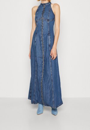 Robe longue en denim bleu sans manches avec fermeture éclair devant, portée avec des bottines bleues assorties, sur une personne debout devant un fond uni.