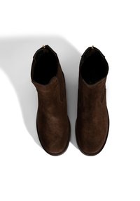 Bottines en suede marron avec un design épuré, présentant des panneaux latéraux élastiques et un détail de fermeture éclair doré à l'arrière. Texture lisse.