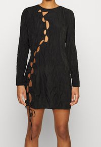 Černé dlouhé šaty s dlouhými rukávy, které mají texturovanou látku, výřezový design na přední straně a šněrování na bocích.