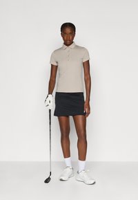 Beige kortærmet polo med krave, parret med en sort skørter. Modellen holder en golfkølle, iført hvide sko og en hvid handske.