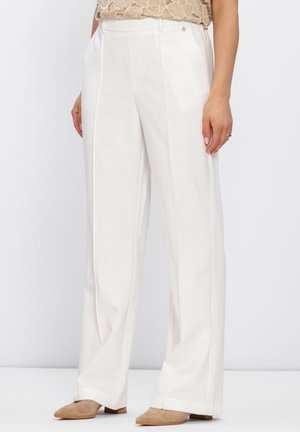 Vrouw met hoge taille, wijde witte broek en beige lage schoenen met puntige neus, staand met handen in de zakken.