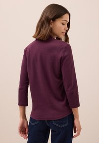 Lila Langarmoberteil mit hohem Kragen, weicher Textur und entspanntem Schnitt. Mit Dreiviertelärmeln und abgerundetem Saum, kombiniert mit Jeans.