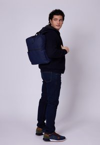 Marinblå ryggsäck med en slät yta och justerbara axelremmar, bärs av en person i en mörk tröja och jeans, med en framficka med dragkedja.