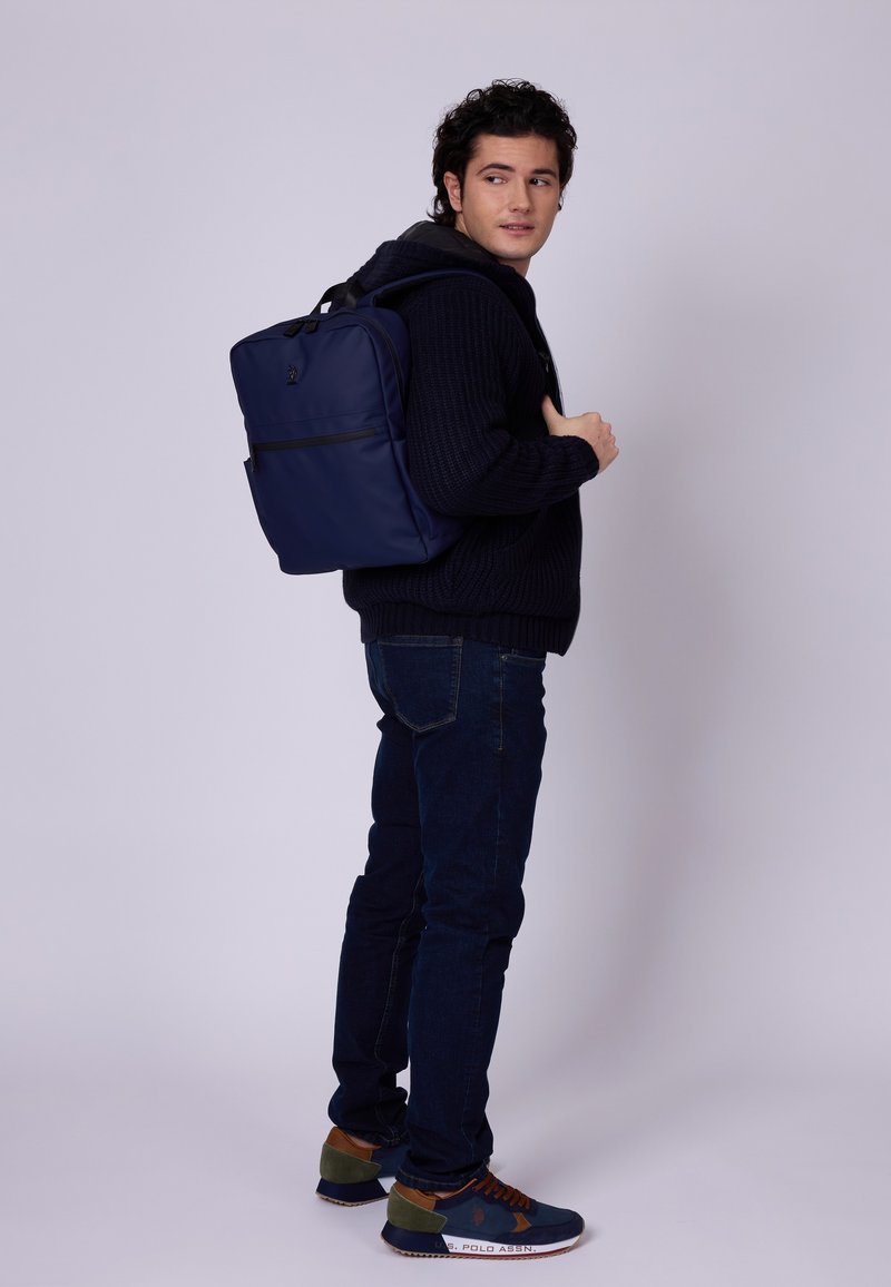 Marinblå ryggsäck med en slät yta och justerbara axelremmar, bärs av en person i en mörk tröja och jeans, med en framficka med dragkedja.