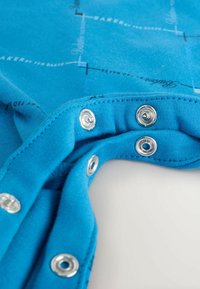 Blaue Baumwollstoffe mit metallischen Druckknöpfen, die ein dezentes Muster mit dem Text "Ted Baker" zeigen. Das Design hat eine glatte Textur und lebendige Farben.