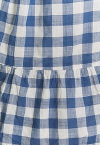 Tissu à carreaux bleu et blanc avec un ourlet froncé. Le matériau semble léger et tissé, présentant des carrés alternés de grande taille dans deux couleurs.