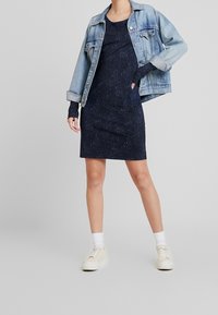 Marinblå klänning med ett subtilt prickigt mönster, kombinerad med en ljus denimjacka. Modellen bär vita sneakers och strumpor.