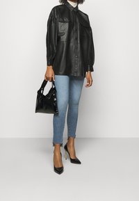 Camisa de cuero negra de gran tamaño con cierre de botones y dos bolsillos en el pecho, combinada con jeans claros deshilachados y zapatos de tacón negros. Sosteniendo un bolso negro.