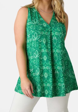 Vrouw draagt een mouwloze groene top met patroon en V-hals, en een witte broek, tegen een effen lichte achtergrond.