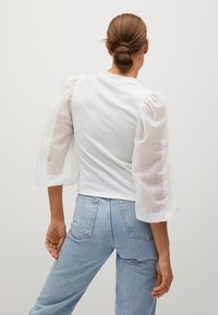 Weißes Bluse mit strukturierten, transparenten Puffärmeln, kombiniert mit hellblauen Jeans. Das Oberteil hat einen runden Halsausschnitt und eine taillierte Passform.