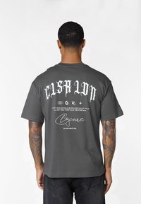 Mann mit tätowierten Armen, trägt ein dunkelgraues T-Shirt mit weißem gotischem Schriftzug und Logos auf dem Rücken vor einem einfarbigen weißen Hintergrund.