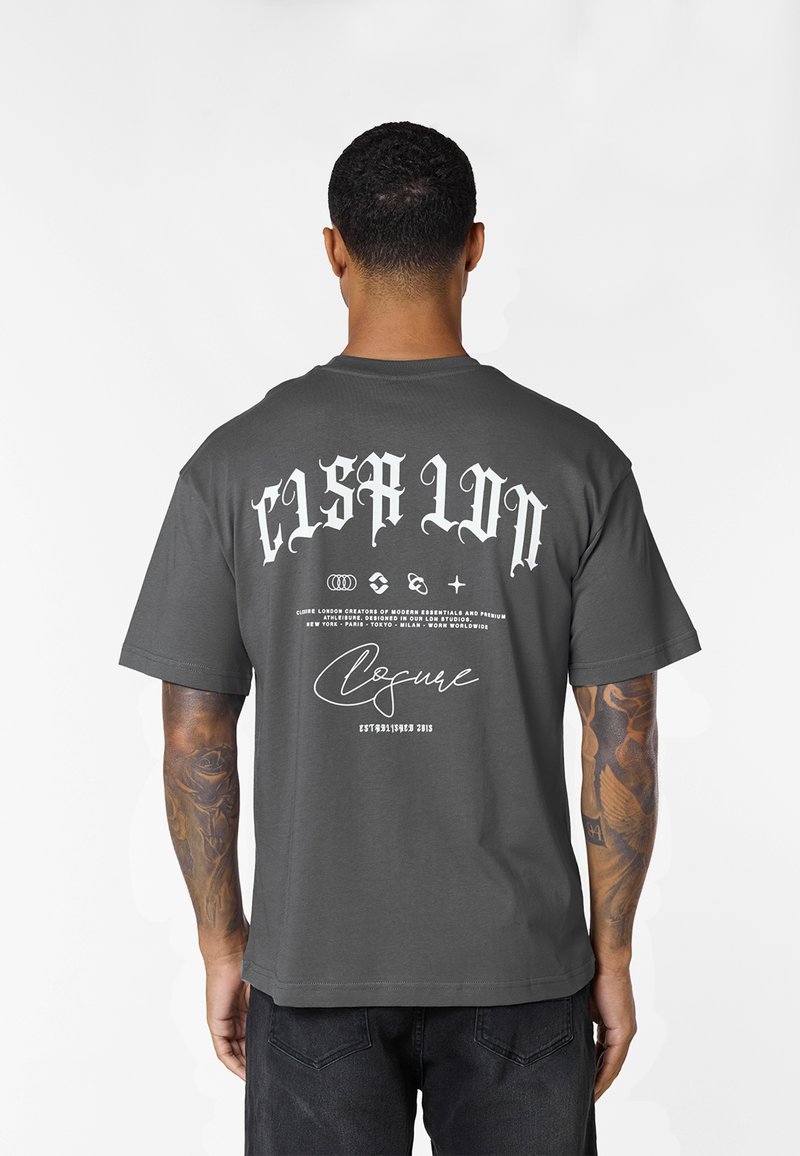 Mann mit tätowierten Armen, trägt ein dunkelgraues T-Shirt mit weißem gotischem Schriftzug und Logos auf dem Rücken vor einem einfarbigen weißen Hintergrund.