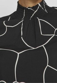 Černý top z látky s vysokým skládaným límcem, s kontrastním bílým abstraktním vzorem a texturovanými detaily podél výstřihu.