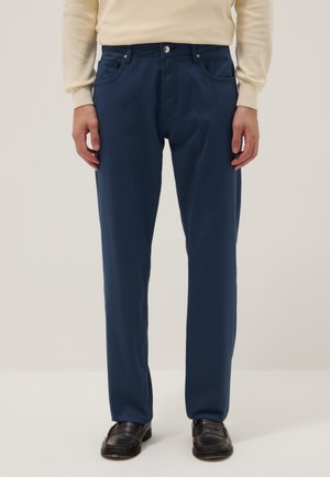 Mann, der dunkelblaue gerade geschnittene Hose, cremefarbenen Pullover und schwarze Leder-Loafer trägt, auf hellem Hintergrund stehend.