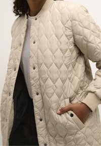 Pehmeäbeige tikattu bomber-takki, jossa resorit hihansuissa ja pääntiessä. Takissa on painonapit ja sivutaskut; sisäpuolella on pilkullinen vuori.