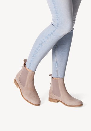 Beine, die hellblaue Skinny-Jeans und beigefarbene Chelsea-Stiefel aus Wildleder mit flachen Holzabsätzen auf einfarbigem Hintergrund tragen.