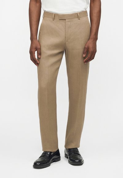 Pantalon beige sur mesure avec pli devant, passants de ceinture, porté avec des mocassins noirs en cuir et une chemise blanche rentrée.